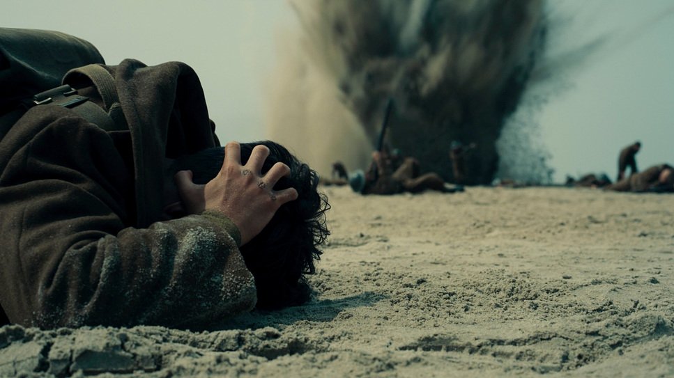 Film still from Dunkirk (2017) showing a young soldier shielding his head as he lies on the ground in front of an exploding bomb