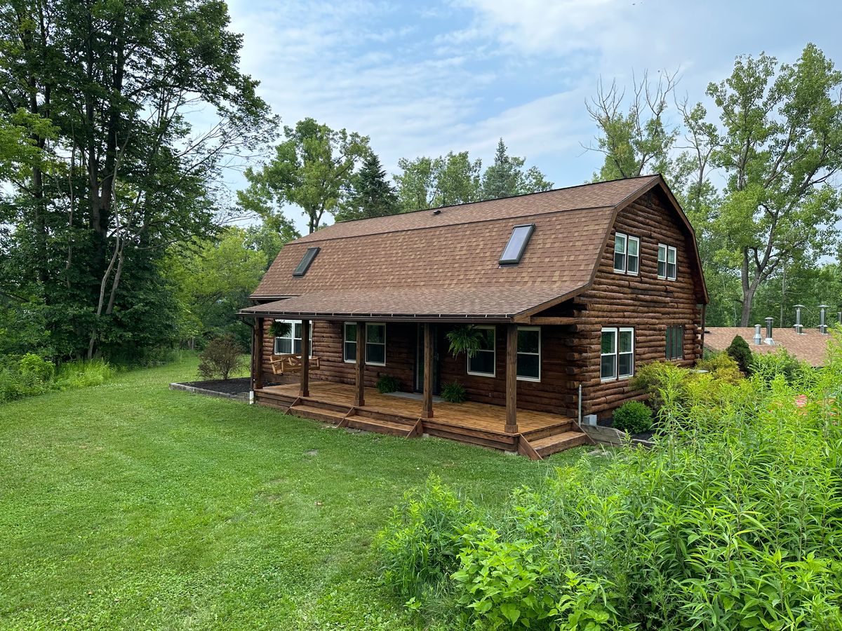 Rustic log cabin with gambrel roof and wrap-around porch surrounded by woodland in upstate New York
