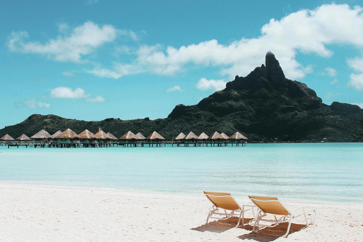 Image of a white sand beach with huts and mountains in the background
