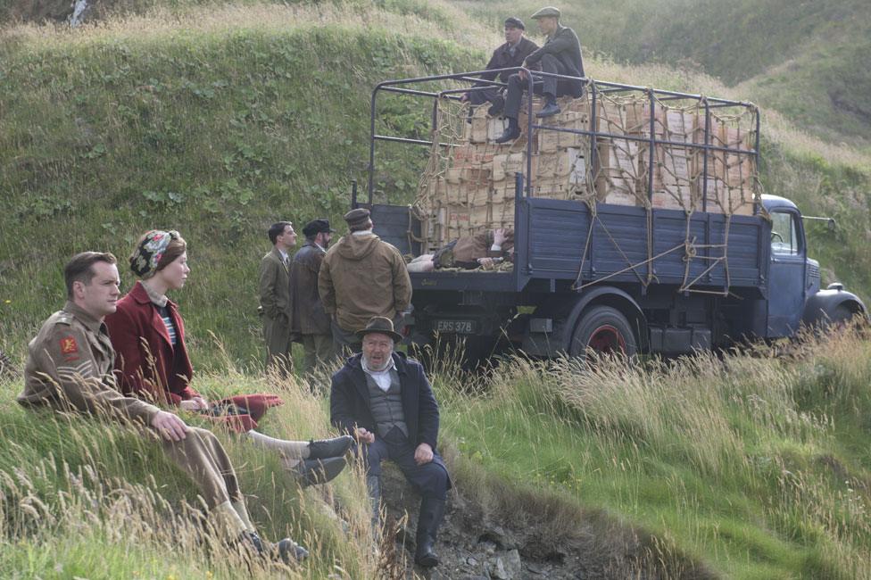 Film still from Whisky Galore! (2016) showing characters sitting on a grassy hilltop.
